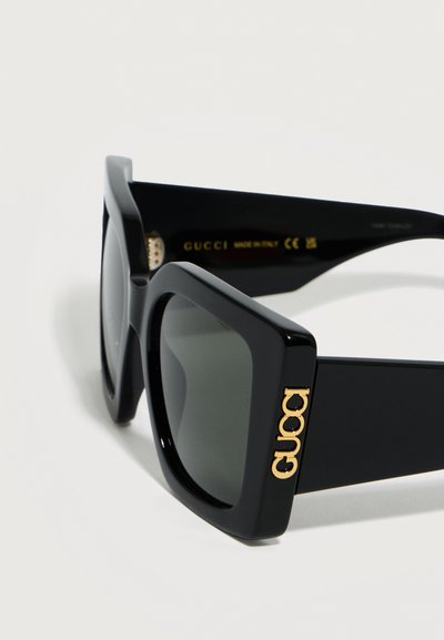 Μεγάλες τετράγωνες μαύρες γυαλιά ηλίου με σκούρους φακούς και χρυσό λογότυπο Gucci στο πλαϊνό μέρος, κατασκευασμένα στην Ιταλία.