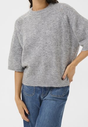 Kvinde iført en lysegrå strikket sweater med et diskret bladmotiv og medium blå denimjeans, poserer med den ene hånd på hoften.