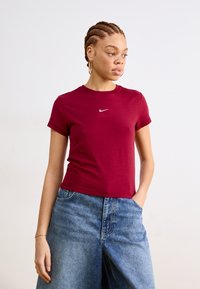 Burgundinė trumpa rankovė marškinė su apvalia iškirpte ir baltu Nike logotipu ant krūtinės, derinami su plačiais mėlynais džinsiniais šortais.