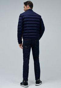 Mann, der eine dunkelblaue steppjacke, dunkelblaue jeans und schwarze sneakers trägt, steht und schaut von einem einfarbigen grauen Hintergrund weg.