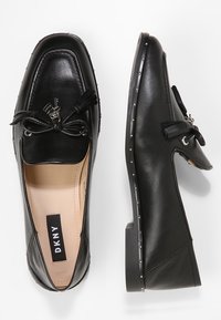 Mocassins en cuir noir avec un orteil carré, un accent en nœud et des éléments décoratifs en métal. Ils sont dotés d'une semelle plate avec un détail clouté le long du bord.