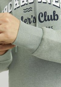 Felpa grigia chiara con polsini a coste e cappuccio con cordino, con scritte in grassetto blu navy che recitano "Jackson's Club" sulla parte anteriore.