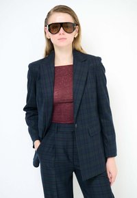 Blazer a quadri blu navy e verde, vestibilità oversize con spalle strutturate. Top aderente bordeaux, pantaloni a quadri abbinati e grandi occhiali da sole scuri.