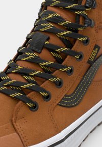 Tummanruskea mokkanahkainen puolikorkea tennari, jossa on mustat ja keltaiset laikukkaat nauhat, näkyvä Vans-logo ja valkoinen kumipohja. Musta sivutrendiviiva.