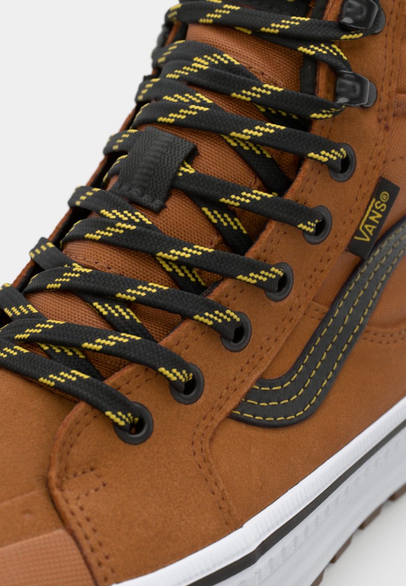 Zapatilla alta de ante marrón con cordones salpicados en negro y amarillo, logo prominente de Vans y suela de goma blanca. Detalle de franja lateral negra.