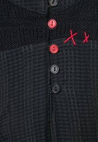 Vêtement texturé noir comprenant un mélange de matériaux tricotés et tissés. Cinq boutons, dont deux rouges et trois noirs, avec des accents brodés rouges.