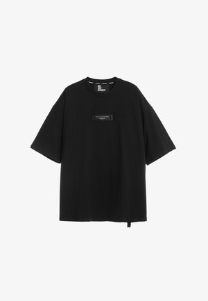 Camiseta negra oversize de manga corta con cuello redondo, pequeño parche rectangular con texto en el pecho y una etiqueta de tela colgando del dobladillo.