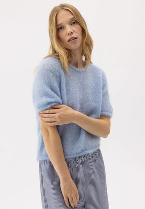 Vrouw met blond haar die een lichtblauwe, pluizige korte mouw trui en gestreepte broek draagt, haar linkerarm aanraakt tegen een effen achtergrond.