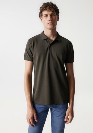 Camisa polo verde oscuro de algodón, con cuello y mangas cortas. Corte ajustado con un logo sutil en el pecho. Jeans de mezclilla claros.