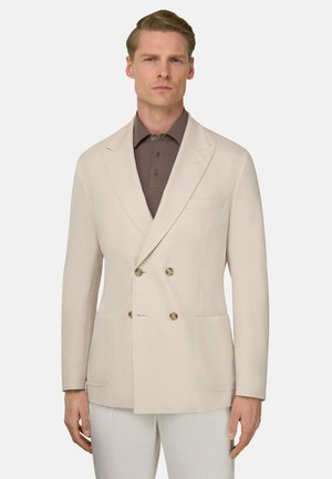 Uomo che indossa un blazer doppiopetto color crema sopra una polo marrone, in piedi contro uno sfondo chiaro e uniforme.