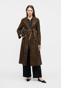 Femme portant un long trench-coat marron ceinturé, un pantalon noir à jambes larges, des ballerines noires pointues, tenant un sac à main noir clouté.