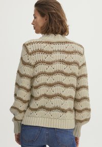 Helles beiges Strickpullover mit Wellen- und Diamantmuster, verziert mit braunen Streifen. Rippbündchen und Saum sorgen für Struktur. Entworfen mit einer lockeren Passform.