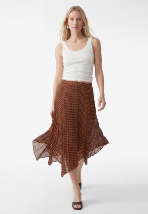 Lyshåret kvinde iført en hvid tanktop, brun plisseret nederdel med prikker, creoløreringe og brune, åbne hæle på en hvid baggrund.