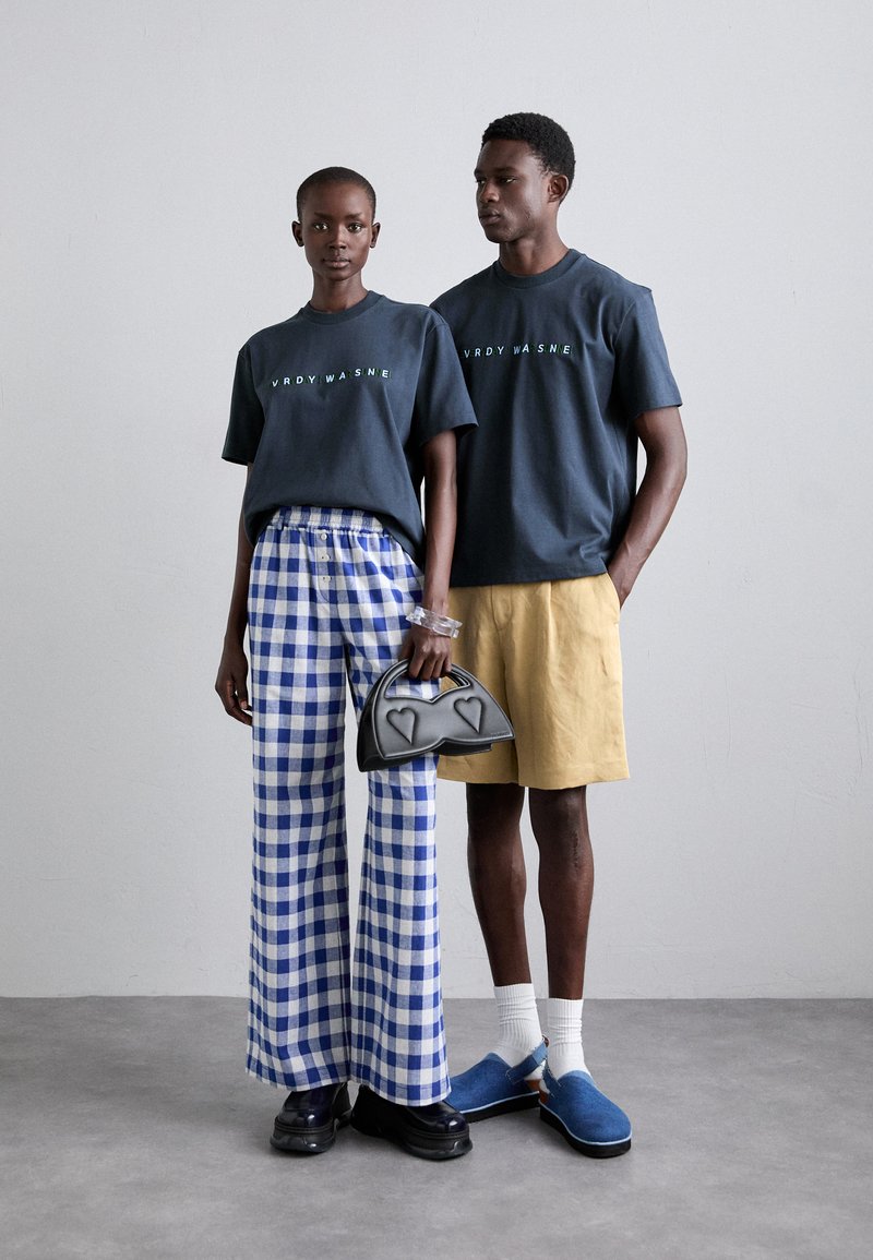 Cotton t-shirts in dark gray with graphic text, paired with blue and white checkered wide-leg pants and khaki shorts. Accessories include a black handbag with heart accents and blue slip-on shoes.