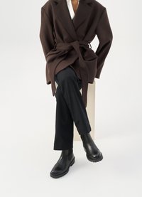 Rjav plašč iz volnene mešanice z pasom, oblečen preko bele srajce in črnih hlač, v kombinaciji s črnimi chelsea škornji, na belem ozadju.