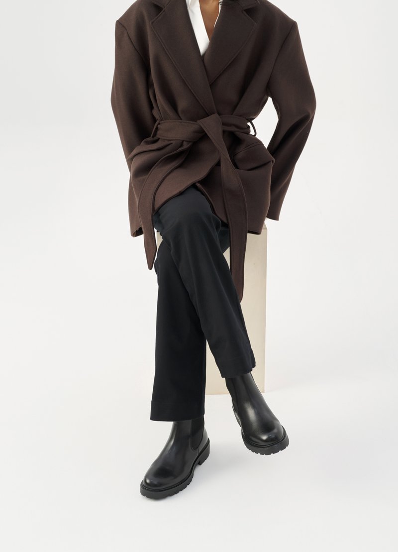 Brun uldblandingsfrakke med bælte, båret over en hvid skjorte og sorte bukser, parret med sorte chelsea støvler, sat mod en hvid baggrund.