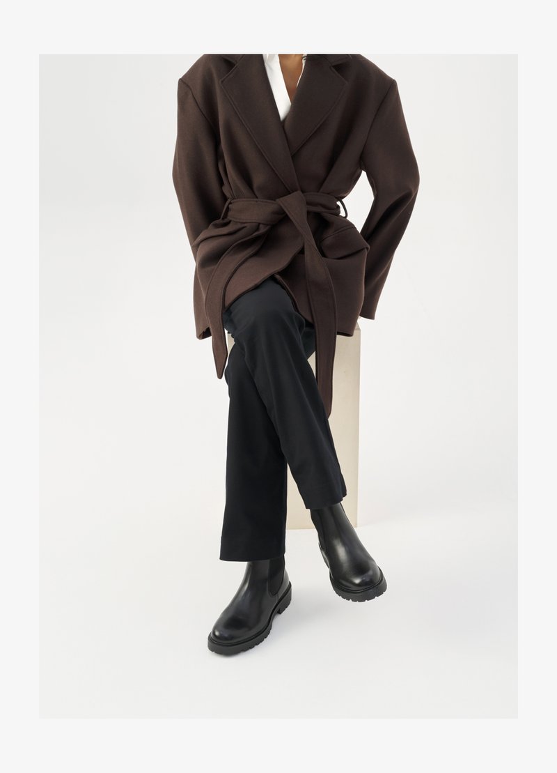 Brun uldblandingsfrakke med bælte, båret over en hvid skjorte og sorte bukser, parret med sorte chelsea støvler, sat mod en hvid baggrund.