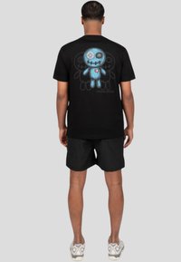 Camiseta de algodón negra con un gráfico de muñeca bordada en azul, con botones y un corazón en la parte posterior, combinada con pantalones cortos negros.
