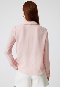Langärmlige Bluse aus weichem pinkfarbenem Stoff. Verfügt über einen Kragen, Knopfmanschetten und einen leicht geschwungenen Saum. Glatte Textur und leichtes Design.