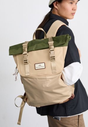 Mochila bege com topo verde, alças ajustáveis e bolso frontal com fecho éclair. Apresenta um logo em patch e detalhe de fecho com cordão.