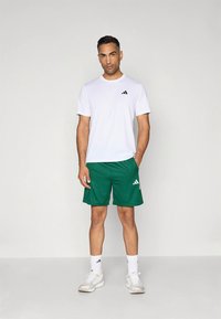 Λευκό T-shirt με κοντά μανίκια και μαύρο λογότυπο, συνδυασμένο με πράσινα αθλητικά σορτσάκια με υφή από δίχτυ, και λευκά αθλητικά παπούτσια.