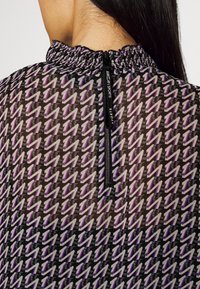 Durchsichtige schwarze Bluse mit Rüschensaum, die ein sich wiederholendes Muster aus lila und grauen Zickzacken aufweist, sowie einem Reißverschluss auf der Rückseite.