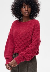 Czerwony sweter dziany z fakturowanym wzorem, oversize'owymi rękawami i okrągłym dekoltem. Tkanina wydaje się miękka i przytulna, zestawiona z ciemnymi spodniami.