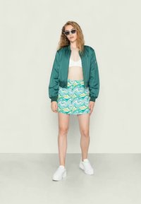 Jaqueta bomber verde sobre um top curto branco, combinada com uma saia mini estampada em espiral teal e amarelo; tênis brancos completam o visual.