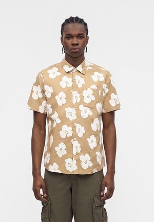 Young man with braided hair wearing a beige short-sleeve button-up shirt with large white flowers and olive cargo pants.