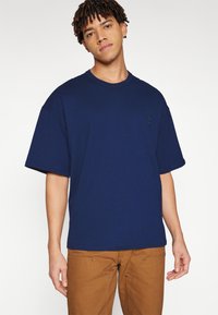 Fiatal férfi rövid, csavart hajjal, laza sötétkék pólót és barna nadrágot viselve, egy egyszerű világos háttér előtt áll.