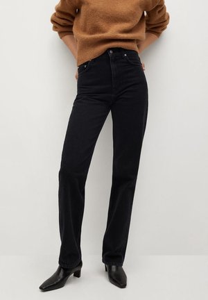 Person trägt schwarze Jeans mit hoher Taille und geradem Bein, schwarze Stiefel mit Absatz und einen braunen Pullover, steht vor einem einfarbigen hellen Hintergrund.