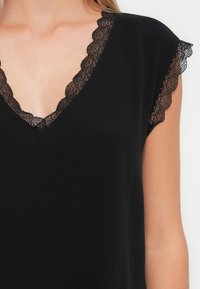 Kvinna iklädd en svart ärmlös topp med V-ringning och spetsdetaljer längs halsringningen och ärmarna mot en enkel bakgrund.