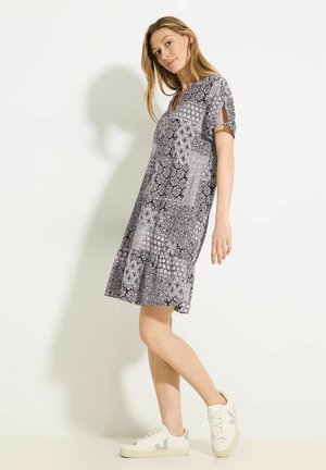 Kleid aus gemustertem Stoff in Schwarz, Weiß und Grau, mit kurzen Ärmeln, einem V-Ausschnitt und einem schwingenden Saum. Kombiniert mit weißen Sneakers.