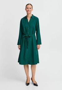 Grünes Hemdblusenkleid mit langen Ärmeln, Taillengürtel und leicht ausgestelltem Saum. Das Material wirkt glatt und hat ein professionelles Design.
