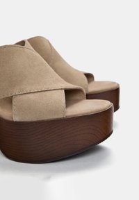 Beige ruskindsplatformsandaler med en buet, mørk træsål og åbent tådesign. Funktioner crossover-stropper og en glat tekstur.