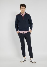 Granatowy sweatshirt z czerwonymi akcentami, z pół-zamkiem, noszony na różową koszulę w kratę. W zestawie z granatowymi spodniami i białymi trampkami.