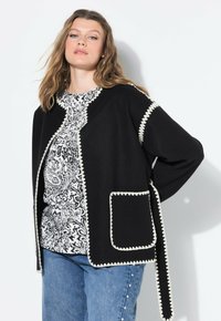 Femme portant un cardigan noir avec des bords scallopés blancs sur un haut à motifs noir et blanc et un jean bleu avec des perles sur les côtés, posant contre un mur blanc.
