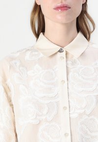 Blusa bianca ricamata con collo, caratterizzata da motivi floreali, una chiusura frontale a bottoni e un accento decorativo di bottone argentato.