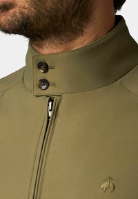 Olijfgroene jas met een hoge kraag, voorzien van twee bruine knopen, een ritssluiting aan de voorkant en een geborduurd logo op de borst. Gladde textuur.
