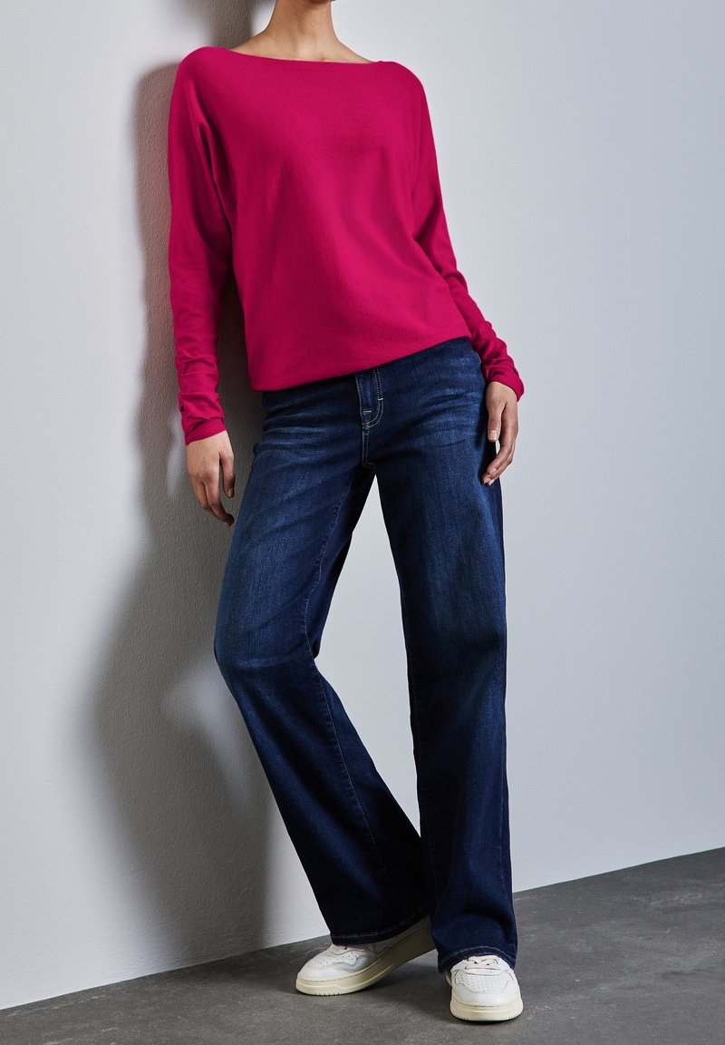 Personne appuyée contre un mur portant une chemise rose vif à manches longues, un jean large bleu foncé et des baskets blanches.