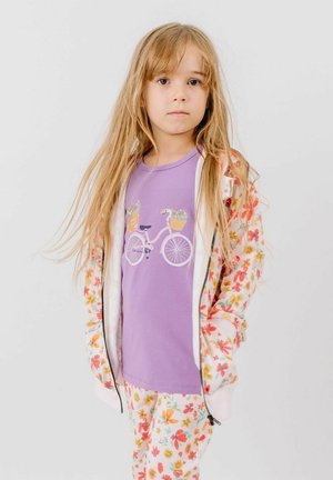 Young girl with long blonde hair wearing a purple bicycle print shirt and white floral jacket and pants, standing against a plain background.