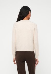 Beige kötött pulóver, kerek nyakkivágással és hosszú ujjakkal, puha textúrával és rövidített fazonnal. Sötétbarna nadrág fölött viselve.