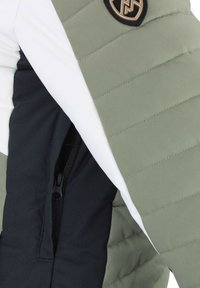 Veste rembourrée avec une palette de couleurs vert olive, blanc et noir. Présente une texture matelassée et une poche zippée. Détail du logo visible.
