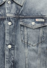 Φωτεινό γαλάζιο τζιν μπουφάν σε κοντινή λήψη που δείχνει μεταλλικά κουμπιά τύπου snap, τσέπη στο στήθος με καπάκι, καφέ ραφές και μια μικρή ετικέτα "CKJK" στο καπάκι της τσέπης.
