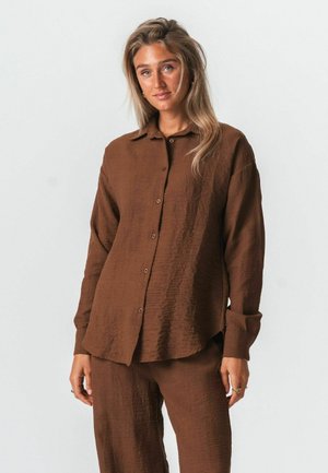 Kvinde med langt blondt hår iført en løs brun tekstureret skjorte med knapper og matchende bukser, stående mod en ensfarvet lys baggrund.