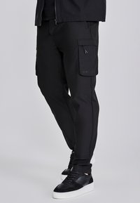 Pantalones cargo negros con un ajuste slim, bolsillos laterales y un logo en la pierna izquierda. Fabricados con un tejido suave y duradero. Combinados con zapatillas negras.