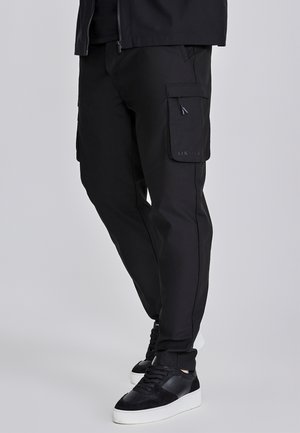Black cargo pants featuring a slim fit, side pockets, and a logo on the left leg. Made of smooth, durable fabric. Paired with black sneakers.