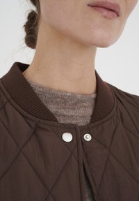 Kvadrajakigt brunt jacka med ribbat krage och silverfärgade tryckknappar, lagd över en ljusbrun texturerad tröja.