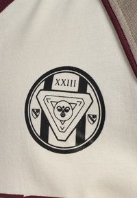 Weißes Stoffstück mit schwarzem, rundem Logo, das ein dreieckiges Design und den Text "XXIII." zeigt. Burgunderfarbene Akzente entlang der Ränder.