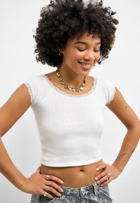 Hvit cropped topp laget av ribbestrikket stoff med et strukturert mønster og blonderkant på ermene og hals Utførelse. Casual passform og myk tekstur.